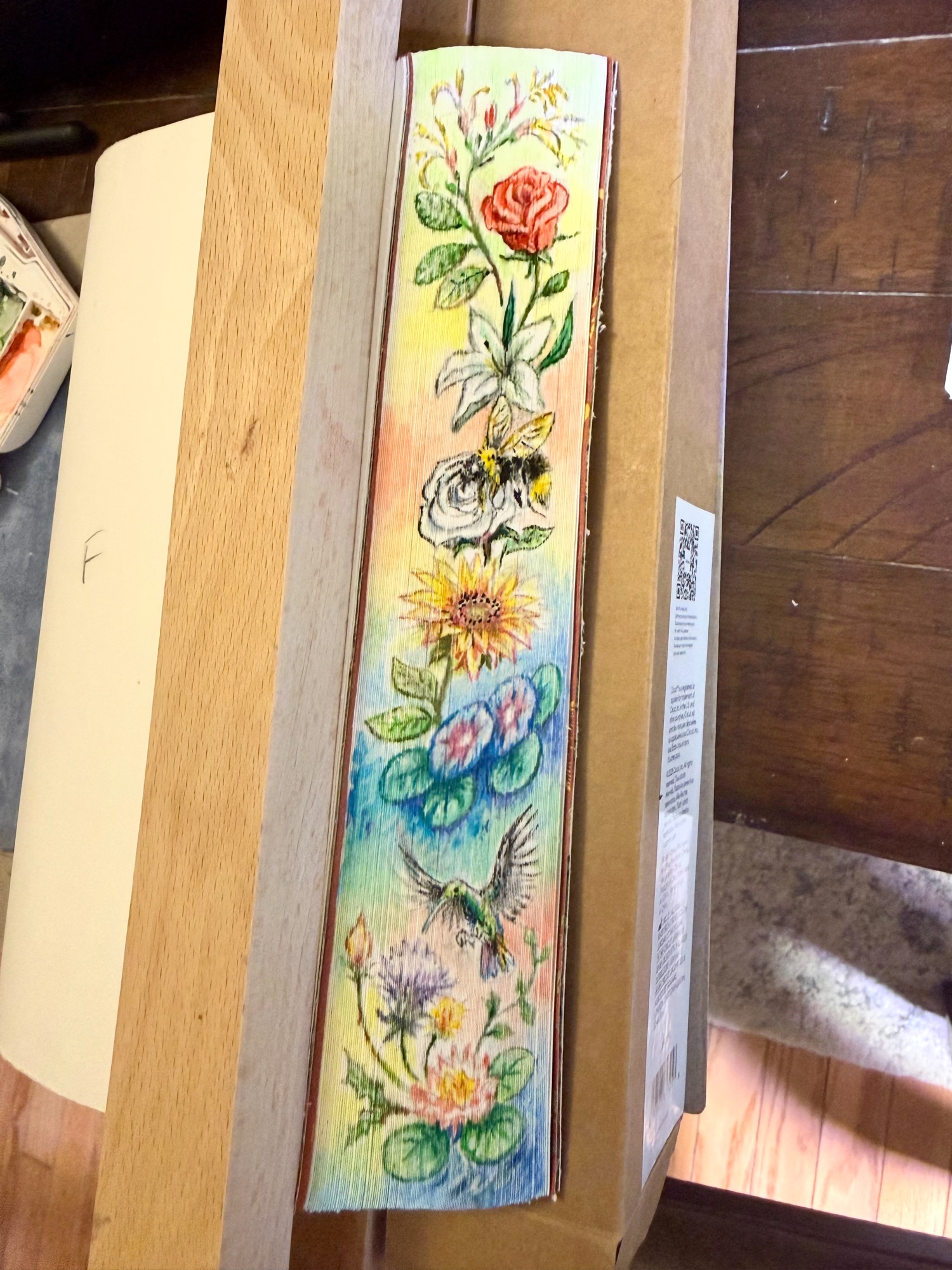 The fore-edge painting of the hand-bound edition of Ritual, revealed with the text block fanned in the press — a watercolour column on the page edges of a red rose, a white lily, a bumblebee on a white rose, a sunflower, blue and pink blooms among lily pads, a hummingbird, and a pink water lily at the foot