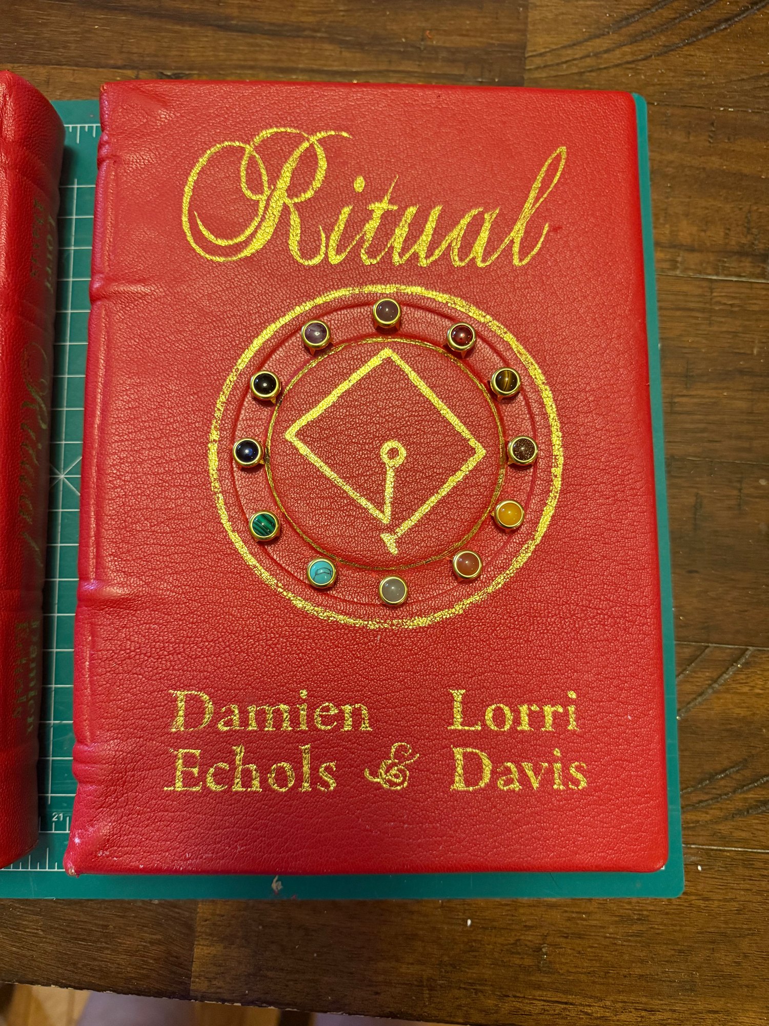 Front board of the hand-bound edition of Ritual — red goatskin, the title tooled in gold script, a ring of cabochon stones in gold bezels around the sigil, and the authors' names tooled below